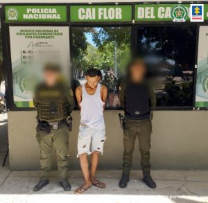 En la imagen está un hombre de pie con la cabeza agachada y los brazos atrás, vestido de camisilla blanca, bermuda de jean azul y sandalias café, custodiado por dos agentes de la Policía Nacional.