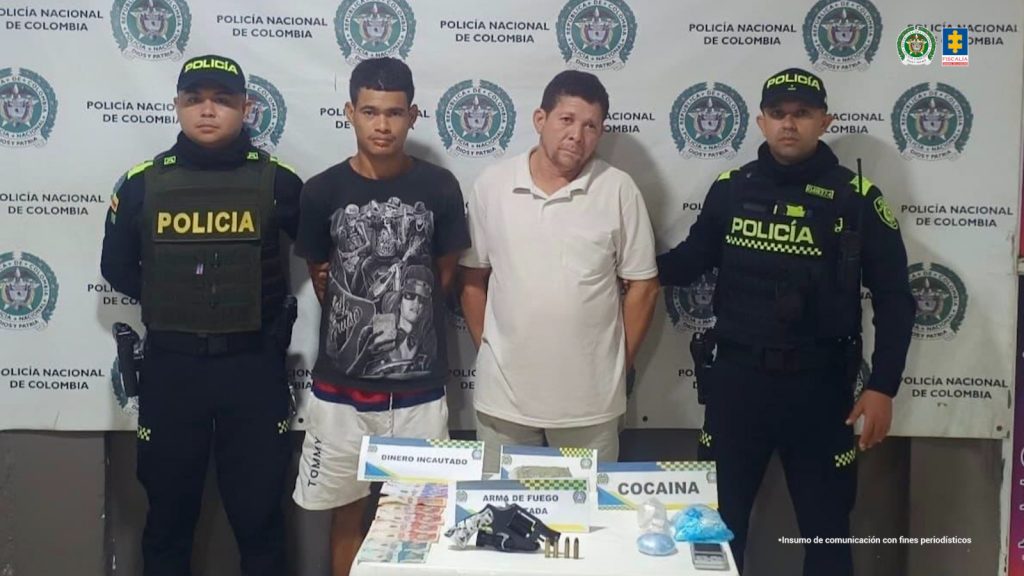 En la fotografía están un hombre joven y otro de mediana edad, trigueños de pie, con los brazos atrás esposados, cabellos negros, uno tiene camiseta negra y el otro camiseta beige con pantalones blancos. Se encuentran custodiados por dos uniformados de la Policía Nacional.