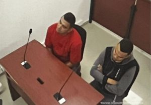 En la fotografía están dos hombres trigueños, con cabellos negros, sentados frente a un escritorio de madera con micrófonos, uno está vestido con camiseta roja y el otro con un buzo gris. Se encuentra en medio de la sala de audiencia donde se les judicializó.