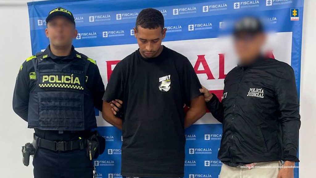 En la imagen está un hombre joven, trigueño, de pie, de frente, con la cabeza agachada, vestido con una camiseta negra y custodiado por un uniformado del CTI y uno de la Policía Nacional. Detrás de ellos hay un pendón que dice Fiscalía. 