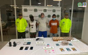 En la imagen aparecen una mujer y dos hombres de pie con las cabezas agachadas y los brazos atrás. Ella está vestida de pantaloneta verde con camiseta blanca, uno de los hombres con camiseta gris y pantalón azul y el otro con camiseta naranja y jean azul. Se encuentran custodiado por dos agentes de la Policía Nacional. Delante de ellos hay una mesa donde se encuentra la droga, el arma y el radio incautado.