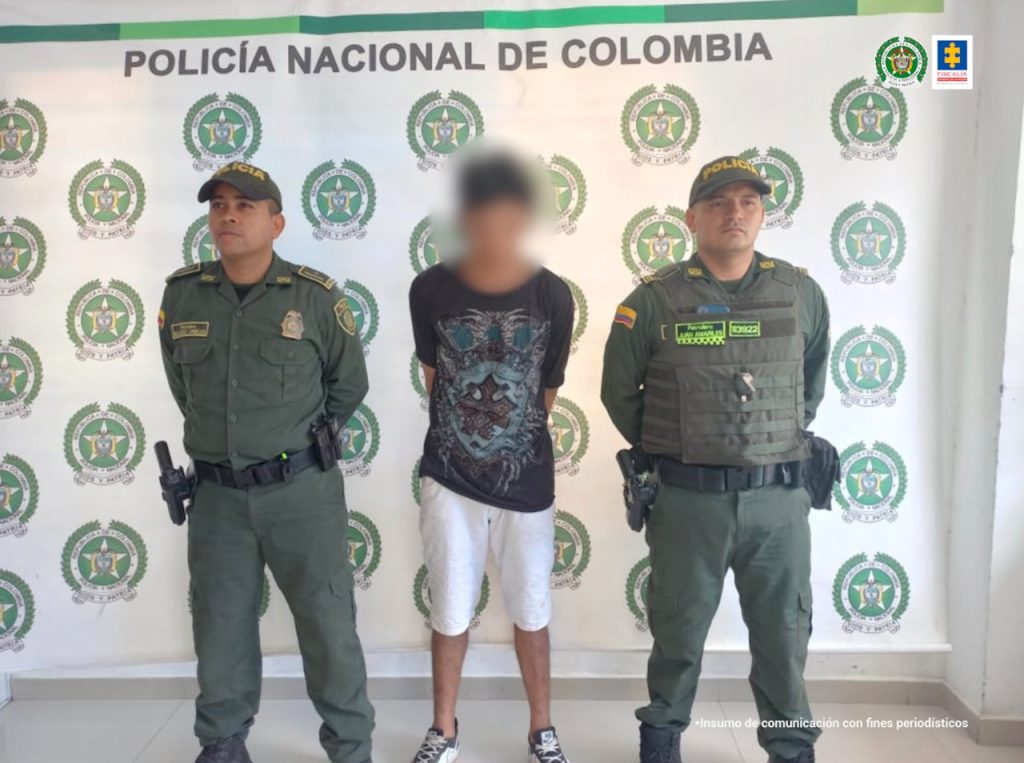 En la imagen se observa a un hombre de bermuda blanca y camiseta negra manga corta, custodiado por dos agentes de la Policía Nacional, delante de un pendón de esa institución.