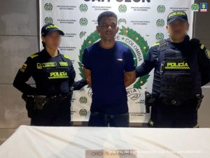 En la fotografía, está un hombre joven, de pie, de frente, con los brazos atrás esposados, trigueño, de cabellos negros cortos, vestido con una camiseta azul y jean azul. Se encuentra custodiado por dos uniformados de la Policía Nacional.