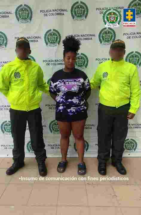 En la fotografía está una mujer morena, de pie, de frente, con los brazos atrás esposados, de contextura gruesa, tiene el pelo negro crespo recogido con una moña, se encuentra vestida con un buzo negro hasta la rodilla y unas sandalias negras. Se encuentra custodiada por dos uniformados de la Policía Nacional.