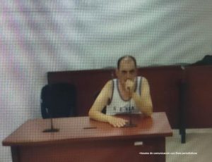 En la fotografía aparece un hombre blanco, semi calvo, con la mano puesta en la boca, vestido con una camisilla blanca y se encuentra sentado en un escritorio de madera con un micrófono, en la sala de audiencia donde se le procesó.