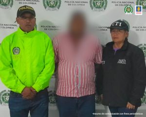 En la imagen se encuentra un hombre de pie, con el rostro difuso camisas de rallas color rojas con blanca, custodiado por un uniformado del CTI y un uniformado de la Policía Nacional.