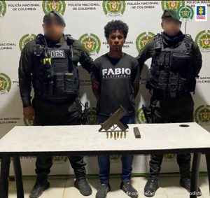 En la fotografía está un hombre de pie, moreno, con los brazos atrás, vestido de camiseta y bermuda negra, custodiado por dos uniformados de la Policía Nacional. Delante de él hay una mesa con el arma que le fue incautada.
