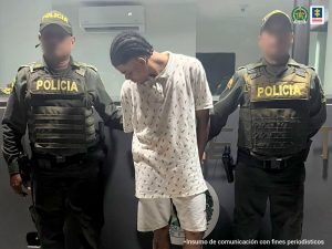 En la fotografía está un hombre de pie, con los brazos atrás, delgado, moreno, vestido de bermuda blanca, camiseta blanca, custodiado por dos agentes de la Policía Nacional.