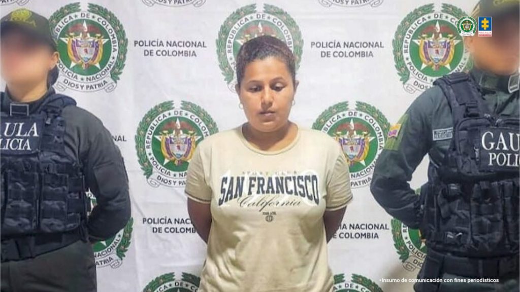 En la fotografía aparece una mujer joven, trigueña de contextura semi gruesa con el cabello recogido, tiene argollas puesta en sus orejas y está vestida con una camiseta amarilla. Se encuentra custodiada por dos uniformados de la Policía Nacional.
