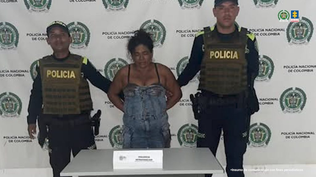 En la fotografía está una mujer de pie, con los brazos atrás esposado, morena, de contextura mediana, vestida con un entrerizo azul, custodiada por dos uniformados de la Policía Nacional.