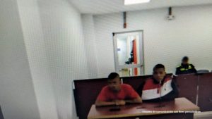 En la fotografía están dos hombres morenos, jóvenes, sentados en un escritorio de madera, con micrófonos, en el interior de una sala asistiendo a las audiencias de control de garantías. 