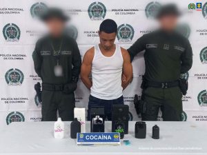 En la imagen aparece un hombre de pie, moreno, con la cabeza agachada, los brazos atrás, vestido de camisilla blanca y jean azul, custodiado por dos uniformados de la Policía Nacional. Delante de ellos hay una mesa con la droga incautada.