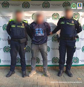 En la fotografía aparece un hombre de pie con las manos atrás, el rostro está difuso y se encuentra vestido con buzo negro, jean gris y tenis. A su lado están dos agentes de la Policía Nacional custodiándolo.