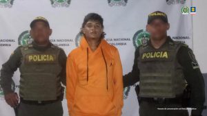 En la imagen aparece un hombre de pie, trigueño, con cabellos castaño larguito, ojos pequeños, boca mediana, vestido con un buzo naranja y se encuentra custodiado por dos uniformados de la Policía Nacional.