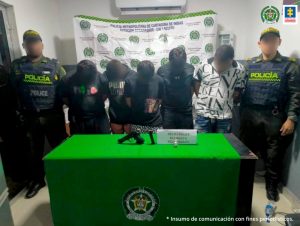 En la fotografía aparecen cinco hombres que se encuentran de pie con las cabezas agachadas, custodiados por dos agentes de la Policía Nacional. Delante de ellos hay una mesa que tiene en la superficie el arma incautada y un reloj.