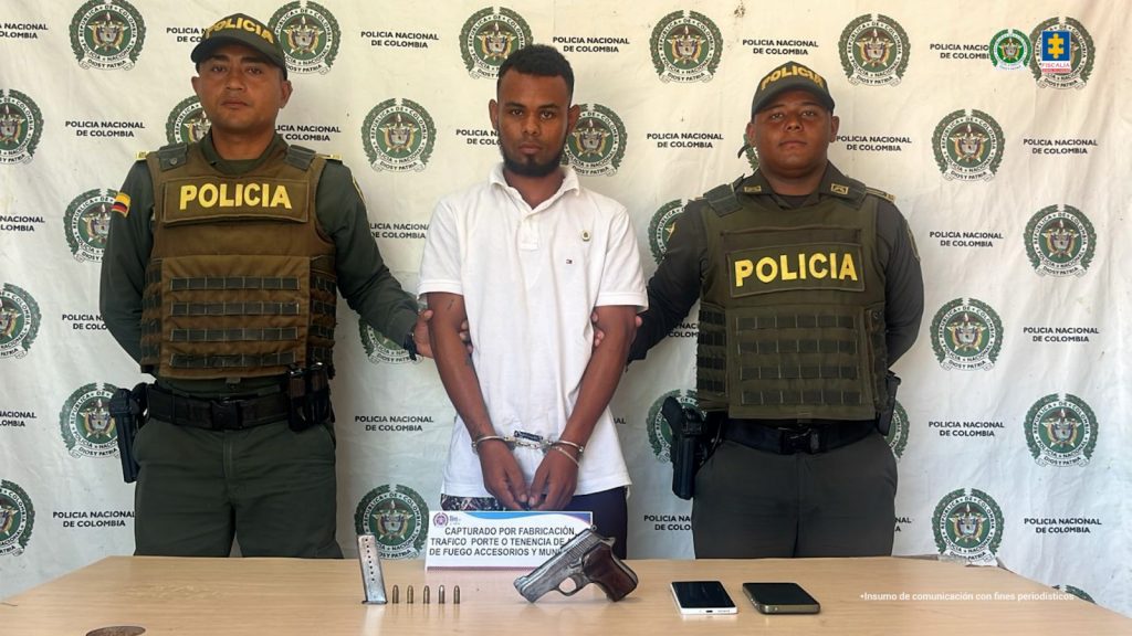 En la fotografía está un hombre joven, de pie, moreno, con los brazos esposados, cabellos y barba negra, vestido con una camiseta blanca y custodiado por dos uniformados de la Policía Nacional.