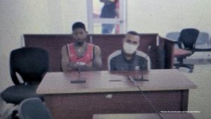 En la imagen están dos hombres morenos sentados frente a un escritorio de madera, uno está vestido con una camisilla roja y el otro con una camiseta azul oscura con rayas blancas y tiene un tapaboca puesto. Se encuentran al interior de la sala de audiencia donde se le procesó.