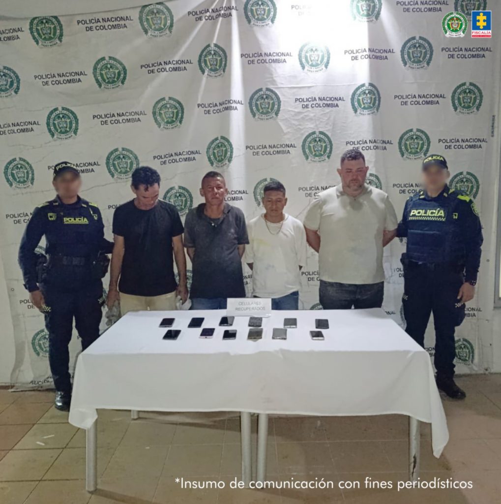 En la foto están cuatro hombres de pie, de frente, custodiado por uniformados de la Policía Nacional. Detrás de ellos hay un pendón de esa institución y delante una mesa con los celulares que les fueron incautados. 