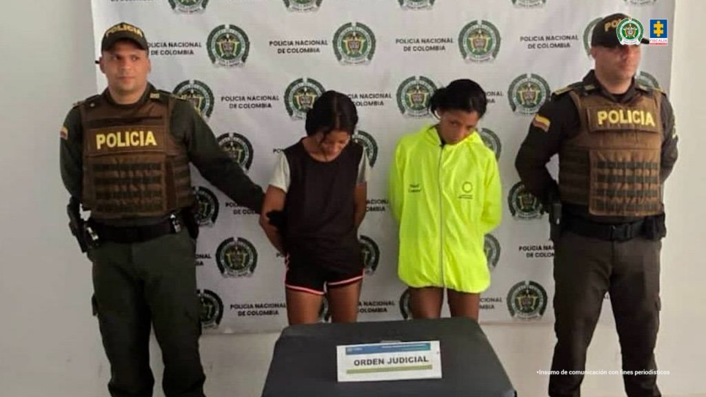 En la fotografía están dos mujeres, gemelas, delgadas, morenas, cabellos negros, de pie, con los brazos atrás esposados, de baja estatura, ambas están vestidas con pantaloneta, una con camiseta negra y la otra con un buzo verde neon. Se encuentran custodiada por dos uniformados de la Policía Nacional.