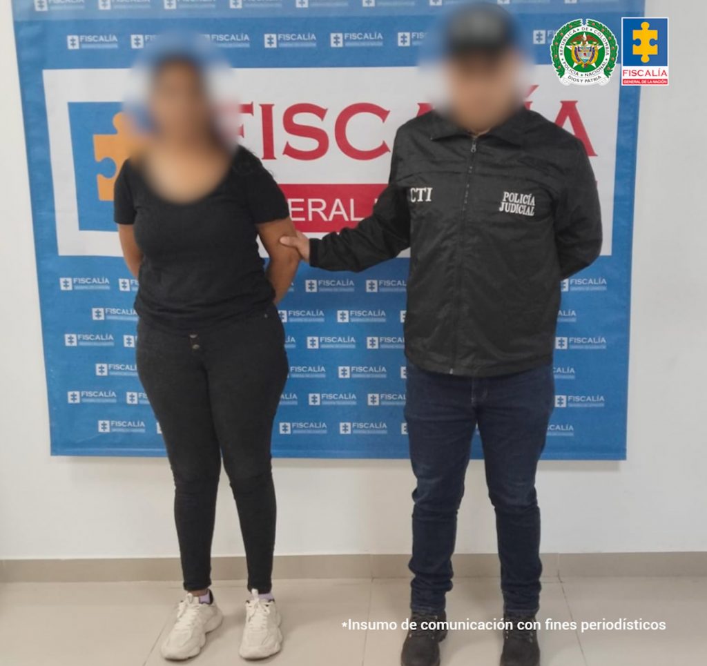 En la fotografía está una mujer de pie con el rostro difuso, los brazos atrás esposados, vestida con una camiseta y jean negros, tennis blanco. Está custodiada por un funcionario del CTI que también tiene el rostro difuso.