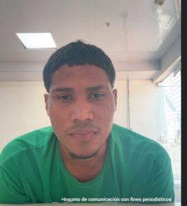 En la imagen está el rostro de un hombre joven moreno de rostro grande ovalado, nariz gruesa, cabellos negros lisos cortos, tiene barba en el mentón. Se encuentra vestido con una camiseta verde, al interior de una sala asistiendo virtualmente a la audiencia en la que se le procesó.