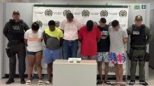 En la imagen, unos hombres y una mujer de pie, con los brazos atrás, custodiados por dos agentes de la Policía Nacional. Detrás de ellos hay un pendón de esa institución.