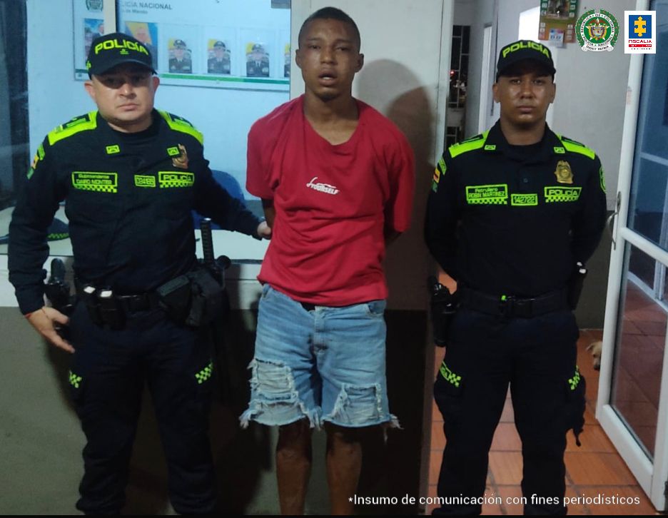  En la fotografía aparece un  hombre moreno de camiseta roja, con bermuda de jean, custodiado por dos agentes de la Policía Nacional en una estación de esa institución.