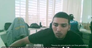 En la imagen está el medio cuerpo de un hombre joven, moreno, cabellos crespos cortos negros, de nariz, orejas y boca grande. Se encuentra vestido con una camiseta negra y está sentado en el interior de una oficina asistiendo virtualmente a la audiencia en la que se le procesó. 