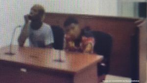 En la fotografía aparecen dos hombres delgados, sentados en frente de un escritorio de madera con micrófonos al interior de la sala de audiencia donde se les procesó.