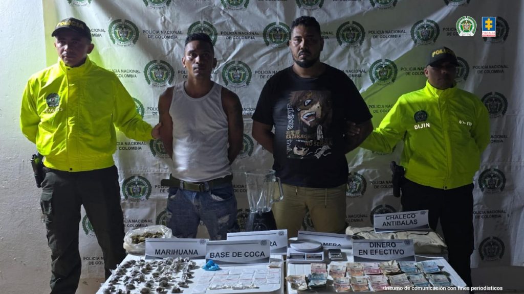 En la imagen están dos hombres de pie, con los brazos atrás esposados, uno es alto y fornido y el otro es de baja estatura. Se encuentran custodiado por dos uniformados de la Policía Nacional. Delante de ellos está lo que se le incautó.