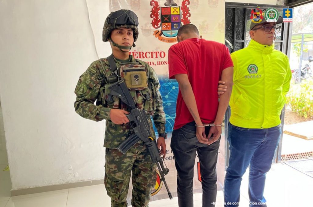 En la imagen está un hombre alto, delgado, está de espalda con los manos esposadas, vestido con una camiseta roja y un jean negro custodiado por uniformados de la Policia y el Ejército Nacional.