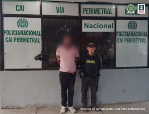 En la fotografía aparece un hombre de pie, moreno, vestido con una camiseta rosada, pantalón negro y tenis blanco, tiene el rostro difuso, con los brazos hacía atrás, custodiado por un uniformado de la Policía Nacional. Detrás de ellos está un aviso que dice CAI vía perimetral.