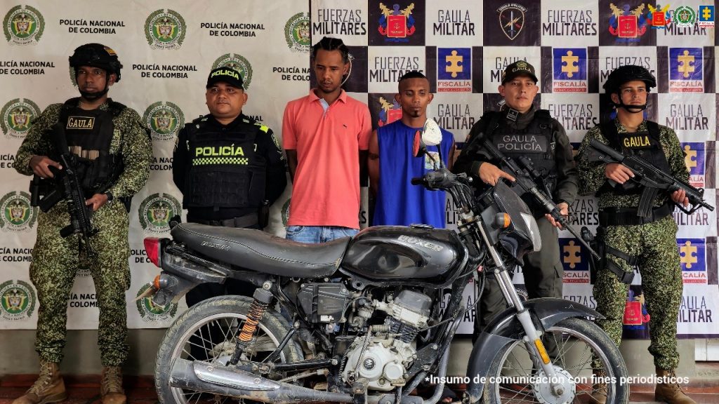 En la fotografía están dos hombres de pie, de frente, con los brazos atrás esposados, morenos de cabellos cortos negros, uno vestido con camiseta rosada y el otro con camiseta azul, custodiados por uniformados de la Policía Nacional y Armada Nacional.