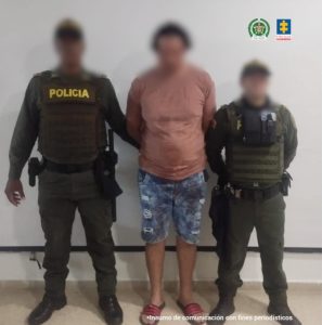En la fotografía está un hombre alto, trigueño, se encuentra de pie, con los brazos atrás, rostro difuso, vestido con una camiseta rosada de bermuda de jean y sandalias rosadas., custodiado por dos uniformados de la Policía Nacional.