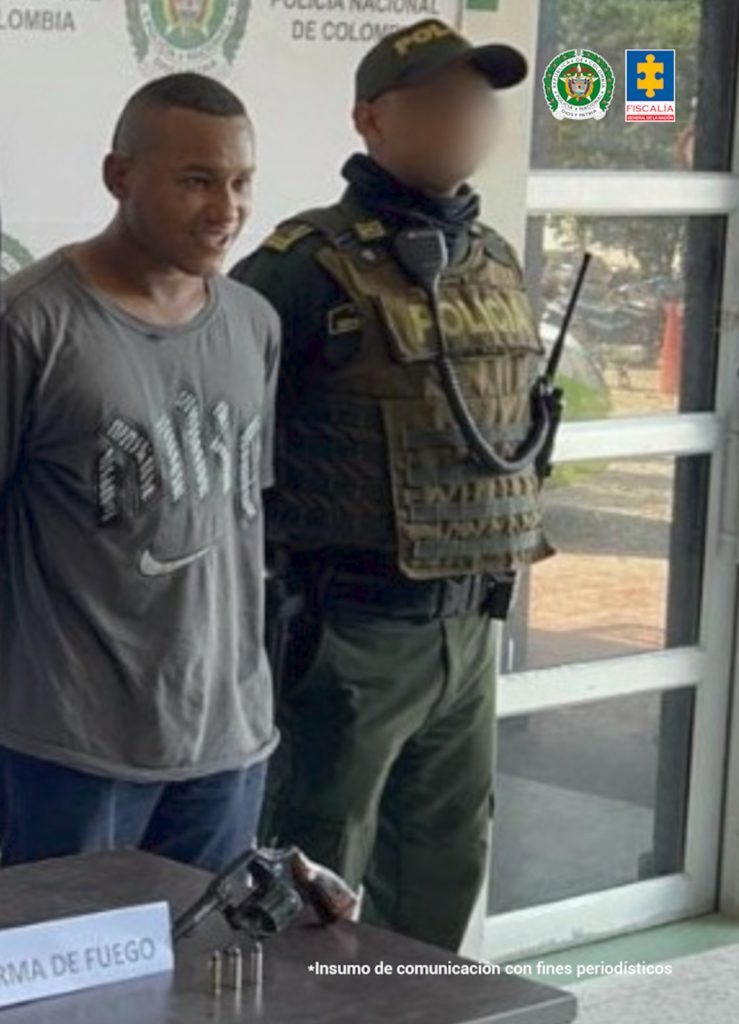 En la imagen aparece un hombre de pie, de estatura baja, moreno, cabellos cortos, vestido de camiseta gris y jean azul, acompañado por un agente de la Policía Nacional. Delante de ellos está el arma incautada.