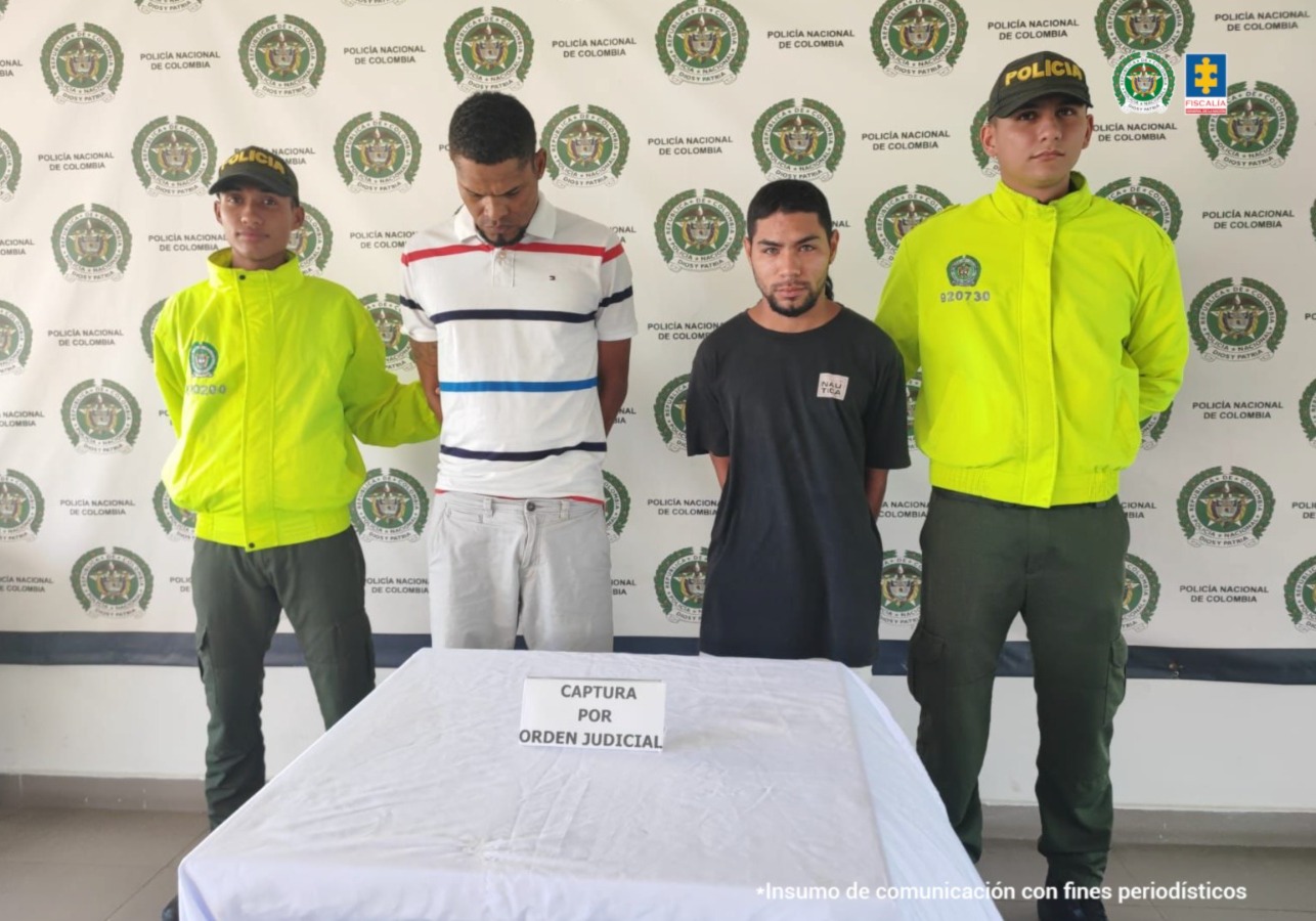 En la imagen aparecen dos hombres de pie, con los brazos atrás esposado, uno es alto moreno delgado y se encuentra vestido con una camiseta blanca con rayas de colores, el otro es de estatura baja trigueño con barba y tiene puesta una camiseta negra. Están custodiado además por dos uniformados de la Policía Nacional.