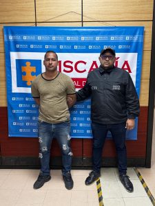 En la fotografía aparece un hombre moreno de pie, de frente, vestido con camiseta café, jean azul y zapatos negros. Se encuentra custodiado por un uniformado del CTI. Detrás de ellos hay un pendón azul de la Fiscalía.