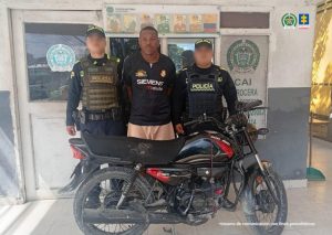 En la foto está un hombe de frente, de pie, con los brazos atrás esposados, moreno, alto, de cabellos cortos crespos, frente grande, vestido con una camiseta negra con pantalón salmón. Se encuentra custodiado por dos uniformados de la Policía Nacional en las afueras de un CAI.