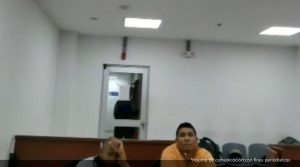 En la fotografía aparecen dos hombres morenos, sentados en un escritorio de madera con un micrófono dentro de una sala de audiencia. Uno se encuentra vestido con camiseta blanca y el otro con camiseta amarilla asistiendo a la audiencia en la que se le procesó.