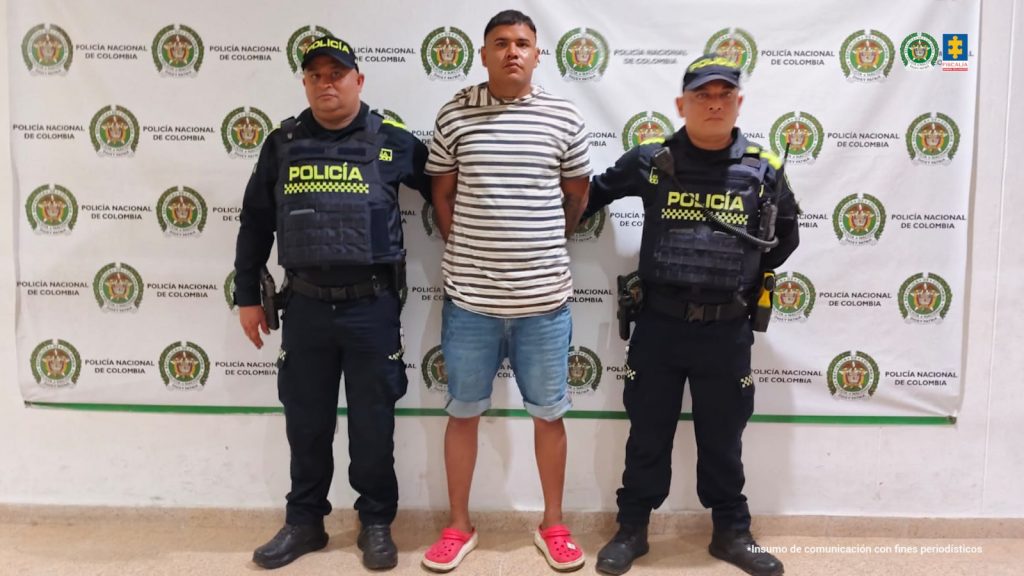 En la fotografía está un hombre trigueño de pie, alto, de frente, de contextura mediana, con los brazos atrás esposados, vestido con una camiseta de rayas negras con blanca, bermuda de jean azul y zapatos rojos. Se encuentra custodiado por dos uniformados de la Policía Nacional.
