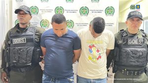 En la fotografía están dos hombres morenos, de pie, de frente, con las cabezas agachadas, los brazos esposados, cabellos negros crespos cortos, uno está vestido con una camiseta azul y el otro con camiseta amarilla. Se encuentran custodiados por dos uniformados de la Policía Nacional.