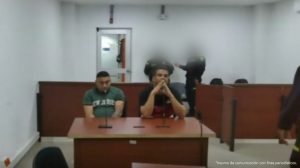 En la imagen están dos hombres sentados en un escritorio de madera con micrófono, uno está vestido con camiseta verde y el otro con camiseta negra, se encuentran acompañados de dos uniformados de la Policía Nacional en la sala de audiencia donde se les procesó.