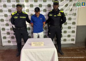 En la fotografía está un hombre blanco, de pie, de frente, con la cabeza agachada, brazos atrás esposados, estatura media, cabellos lisos negros, está vestido con camiseta azul oscura y jean azul claro. Se encuentra custodiado por dos uniformados de la Policía Nacional. 