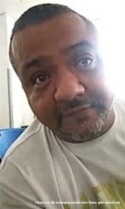 En la fotografía aparece el medio cuerpo de un hombre moreno de barba canosa, ojos negros, semi calvo, nariz y ojos grande, vestido con una camiseta blanca. Se encuentra sentando al interior de una oficina asistiendo virtualmente a la audiencia en la que se le procesó.