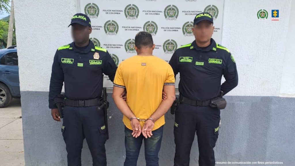 En la fotografía aparece un hombre de pie, se encuentra de espalda con los brazos esposados, vestido de camiseta amarilla y jean azul, custodiado por dos agentes de la Policía Nacional.
