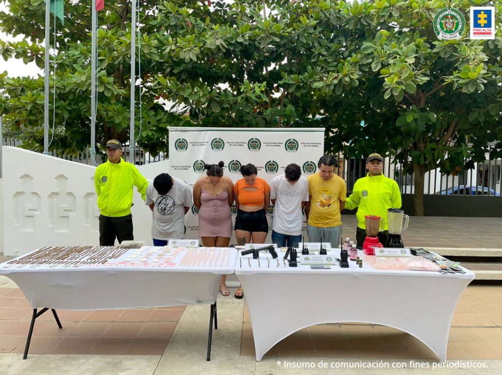 En la imagen se observan a los capturados con la cabeza agachada custodiados por dos agentes de la Policía Nacional delante de un pendón de esa institución. También se divisa una mesa con la droga y las armas incautadas.