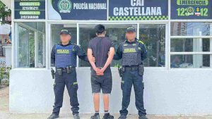 En la fotografía está un hombre blanco, de pie, de espalda con los brazos atrás esposados, de cabellos negros lisos, vestido con camiseta, bermuda, medias y tenis negros. Se encuentra en las afueras de un CAI custodiado por dos uniformados de la Policía Nacional. 