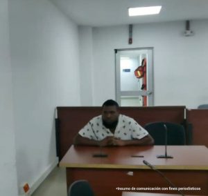 En la fotografía está un hombre moreno de contextura gruesa, cabellos crespos negros, barba negra, se encuentra vestido con una camiseta blanca con negro y está sentado en un escritorio de madera con un micrófono al interior de la sala de audiencia donde se le procesó.