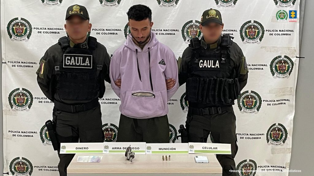 En la fotografía está un hombre de pie con los brazos atrás esposado, tiene el cabello negro, la cabeza agachada, barba y está vestido con un buzo de color lila. Se encuentra custodiado por dos uniformados de la Policía Nacional.