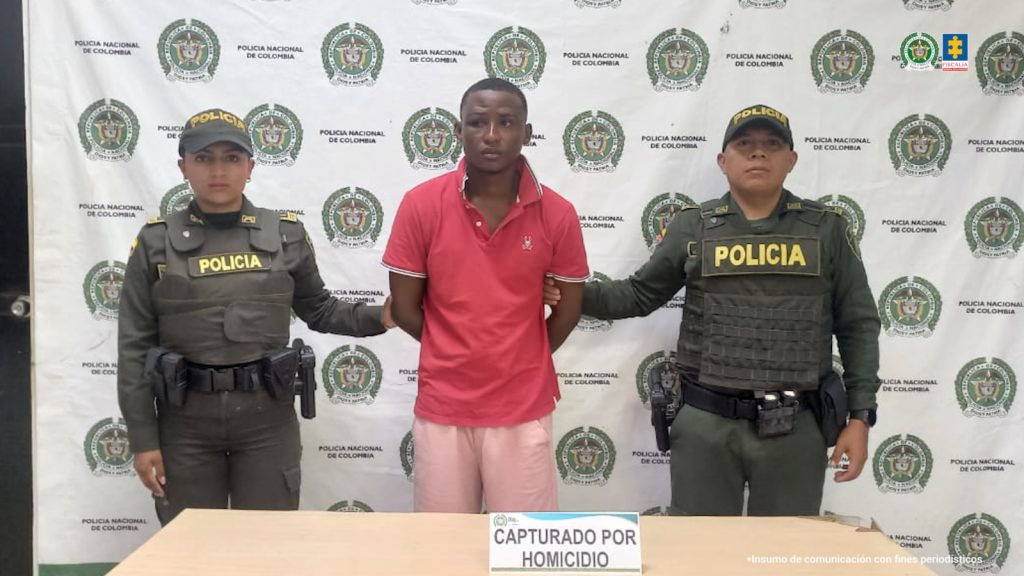 En la fotografía está un hombre de pie, moreno, de cabellos negros crespos, orejas grandes y labios gruesos, vestido con una camiseta roja y una bermuda rosada.Se encuentra custodiado por dos uniformados de la Policía Nacional.